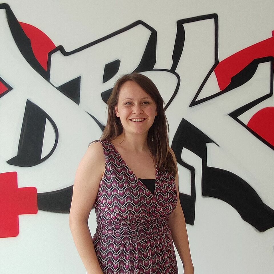 Zu sehen ist Rebecca Lurtz, Bildungsreferentin des Jugendrotkreuzes. Sie trägt eine lila Kleid. Sie steht vor einem Graffiti auf dem DRK steht und ein rotes Kreuz gesprüht ist. Die Wand im Hintergrund ist weiß.