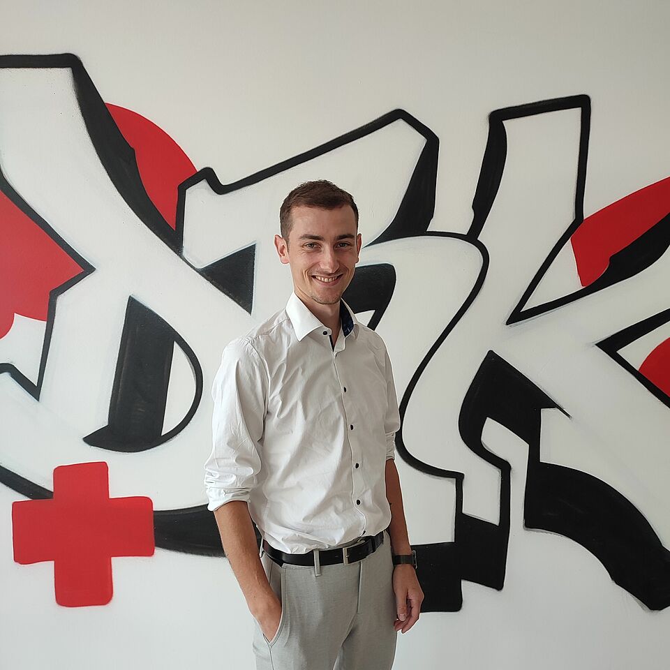Zu sehen ist Giuliano Hess der Landesreferent des Jugendrotkreuzes. Er trägt ein weißes Hemd und eine beige Hose , die Hände stecken in den Hosentaschen. Er steht vor einem Grafitti auf dem DRK steht und ein rotes Kreuz gesprüht ist. Die Wand im Hintergrund ist weiß.