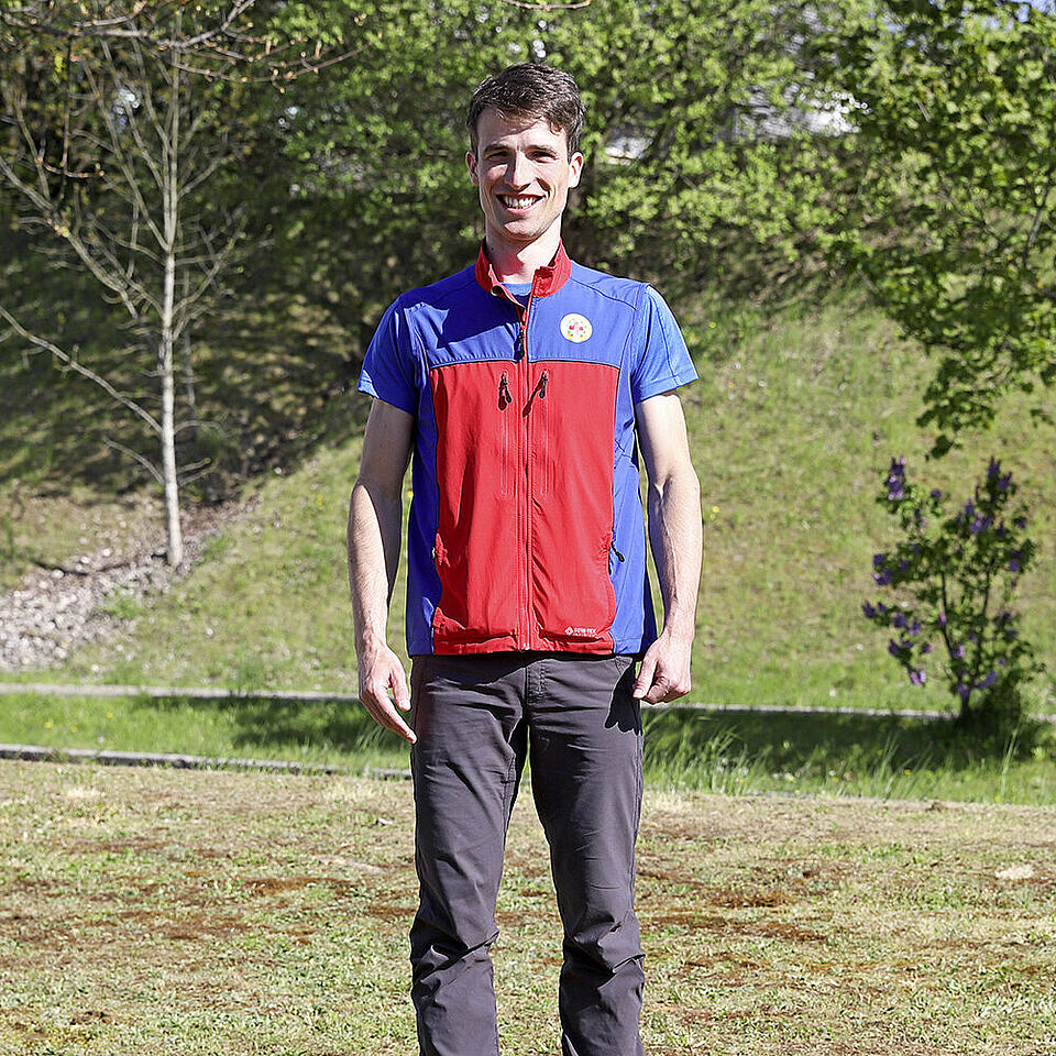 Zu sehen ist Lukas Pfletschinger mit einer Bergwachtweste, die rot und blau ist. Er hat kurze braune Haare und lächelt. Im Hintergrund ist eine Böschung mit Gras und Bäumen zu sehen.