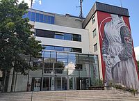 Zu sehen ist ein Gebäude.Es ist die Landesgeschäftsstelle des DRK-Landesverbandes Baden-Württemberg. In der Mitte des Bildes sieht man den gläsernen Eingang. Rechts davon ist ein recht hoher Gebäudeteil. Auf diesem Gebäudeteil ist ein Graffiti. Das Graffiti bildet eine Frau in einer medizinischen Schürze aus dem 19. Jahrhundert ab.