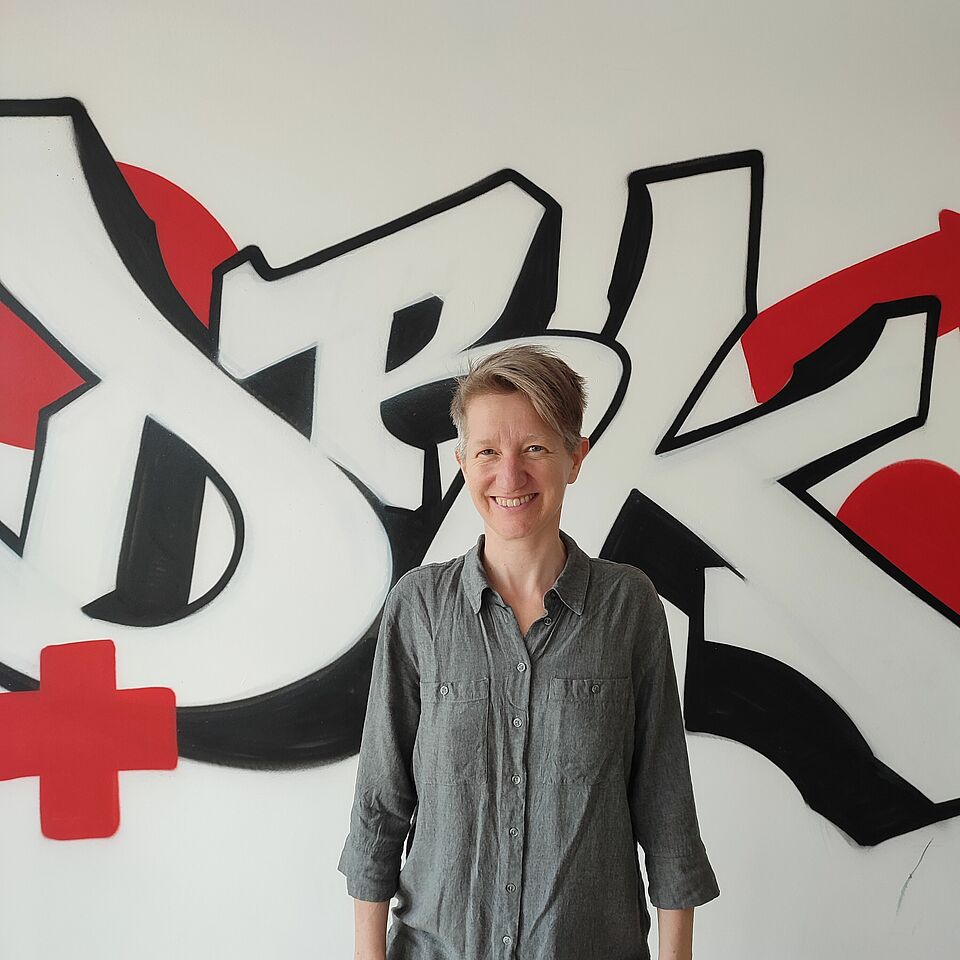 Zu sehen ist Anne Schäfer, Bildungsrefrentin des Jugendrotkreuzes. Sie trägt eine graue Bluse und eine blaue Jeans. Sie steht vor einem Graffiti auf dem DRK steht und ein rotes Kreuz gesprüht ist. Die Wand im Hintergrund ist weiß.